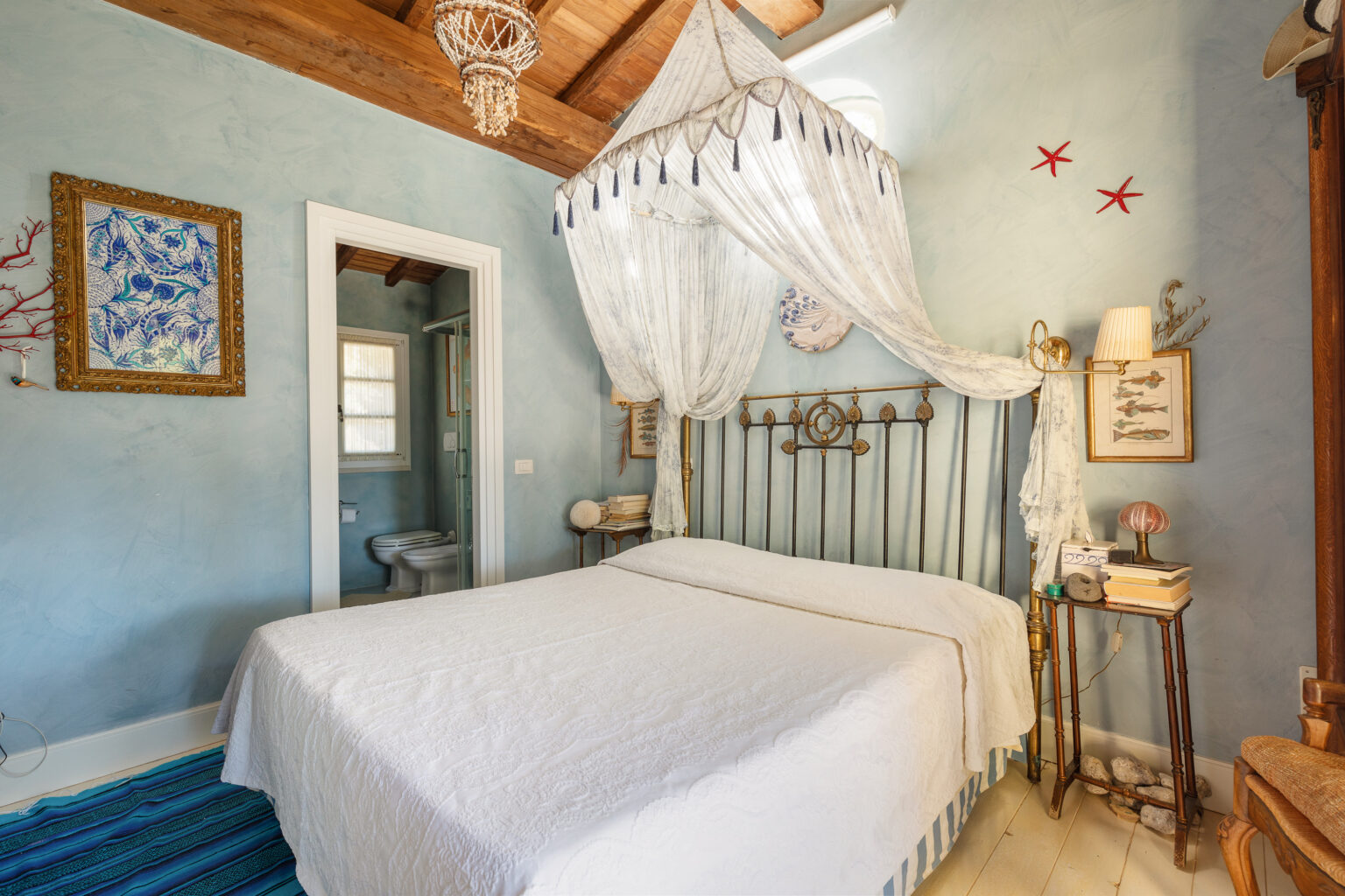Cozy bedroom featuring a wrought iron bed with a white quilt, decorative wall art, and a vintage chandelier. The room has light blue walls, wooden beams, and a small en-suite bathroom visible through an open door. Natural light fills the space, enhancing the inviting atmosphere. | Only on SopranoVillas