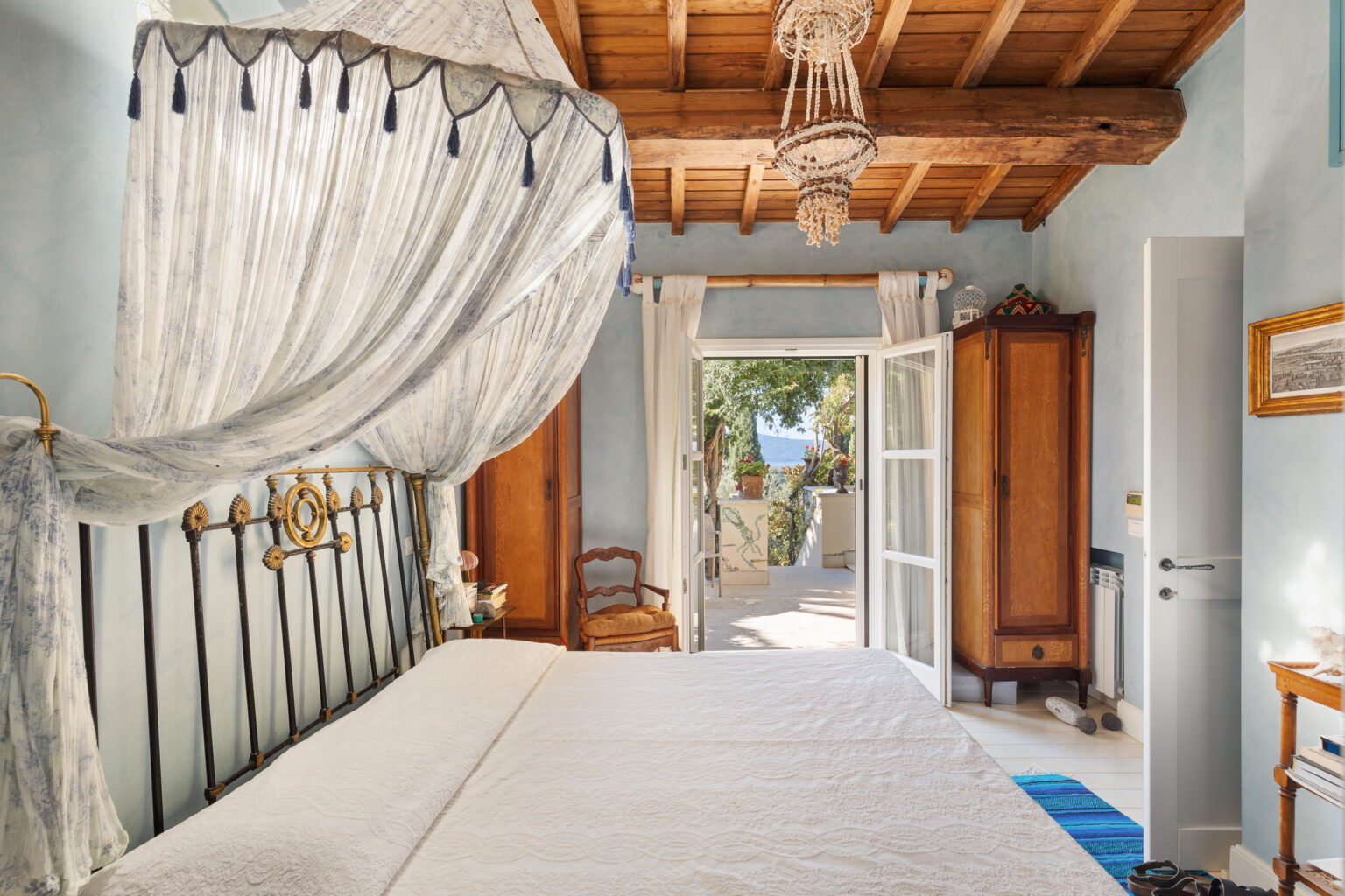 Cozy bedroom featuring a decorative canopy bed with an intricate headboard, soft white bedding, and a charming wooden wardrobe. Natural light floods the room through open French doors, revealing a glimpse of greenery and outdoor space. Elegant details include a beaded chandelier and light blue walls, creating a serene atmosphere. | Only on SopranoVillas