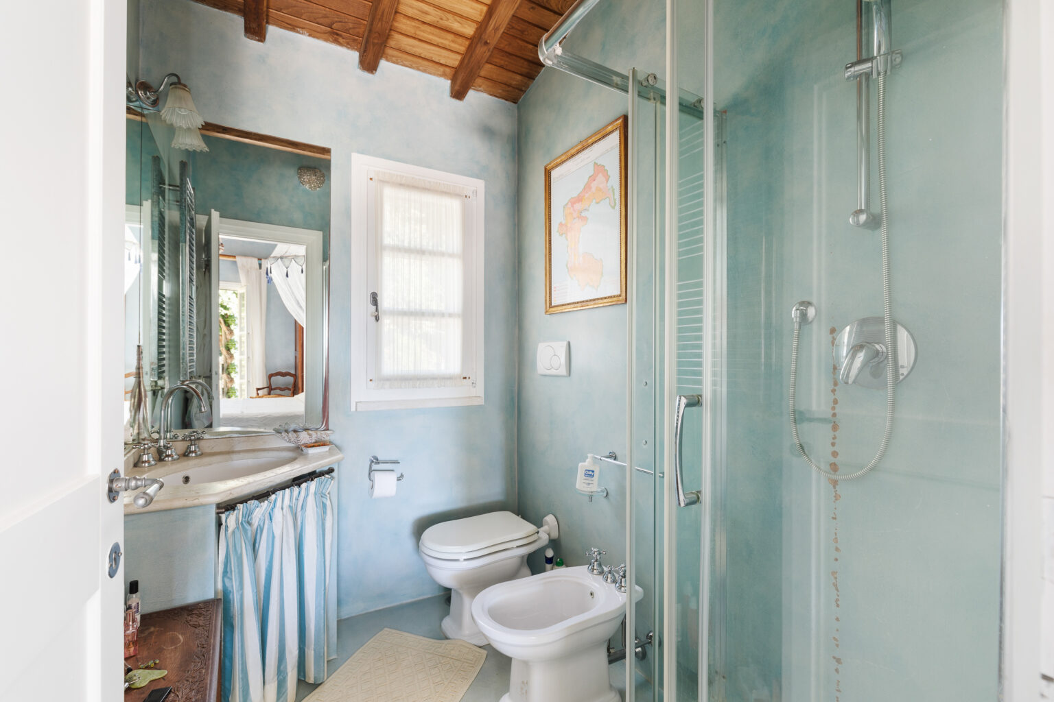 Modern bathroom with blue walls, wooden ceiling, and glass shower. Features a stylish sink with a mirror, a bidet, and a toilet. Natural light streams in through the window, enhancing the serene atmosphere. | Only on SopranoVillas