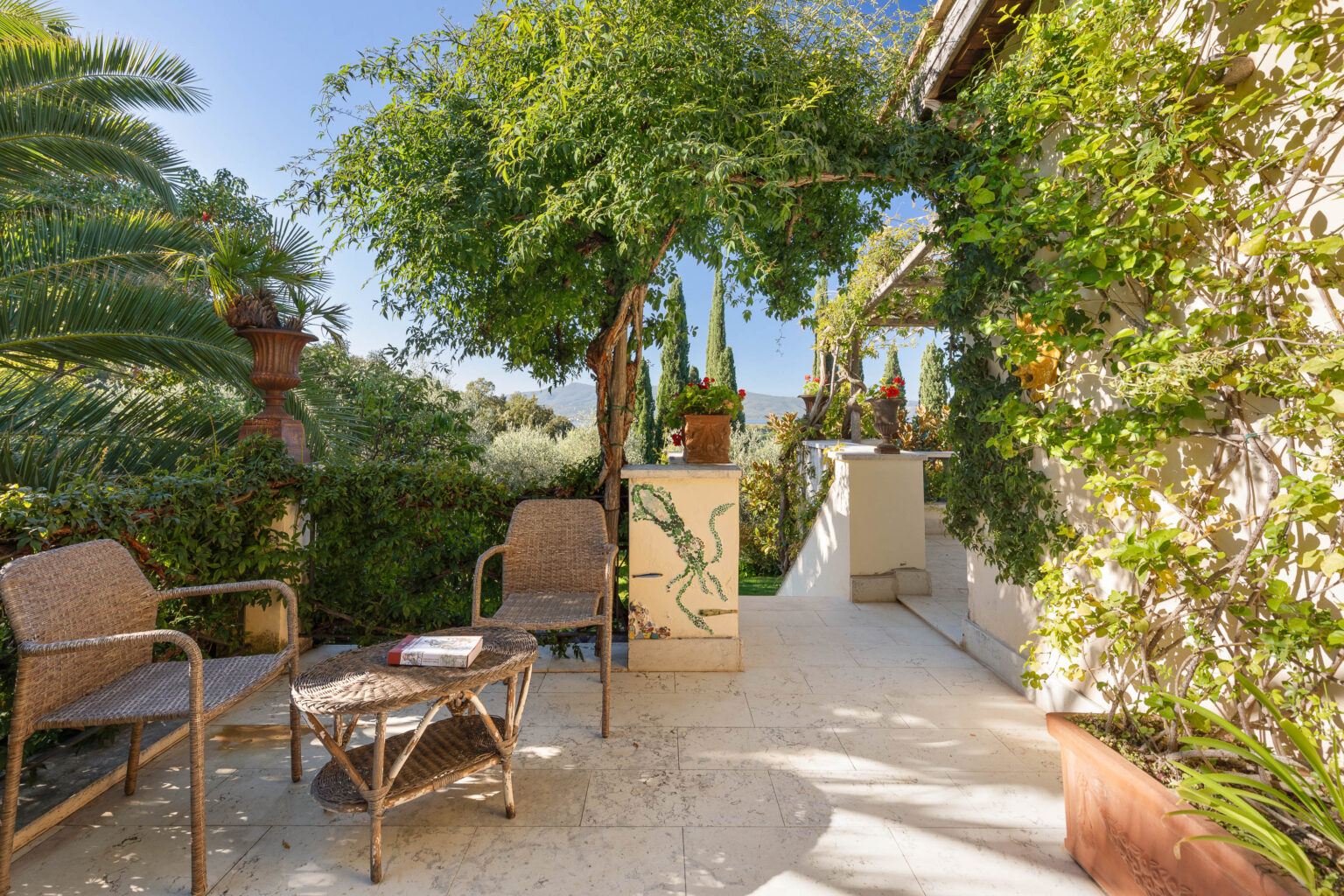Cozy outdoor patio featuring two wicker chairs and a round coffee table, surrounded by lush greenery and vibrant flowers. The scene includes a stone wall with decorative plants and a view of distant mountains under a clear blue sky, creating a serene and inviting atmosphere. | Only on SopranoVillas