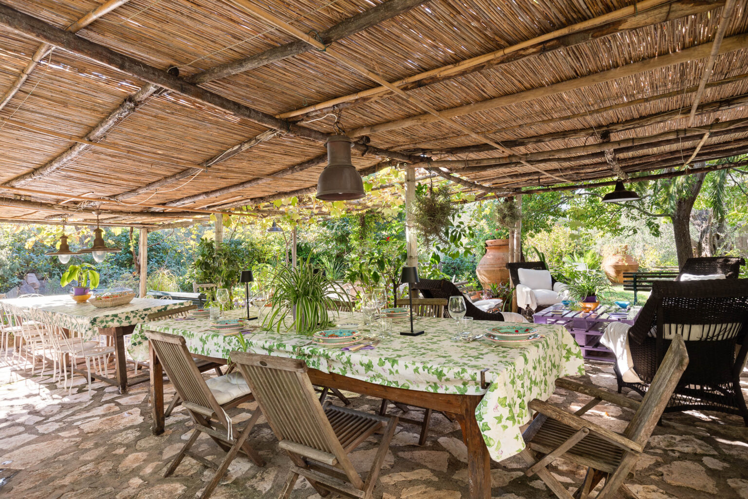 Outdoor dining area featuring a rustic wooden table with a floral tablecloth, surrounded by folding chairs and decorative plants. The space is covered with a thatched roof, illuminated by hanging lights, creating a cozy atmosphere in a lush garden setting. | Only on SopranoVillas