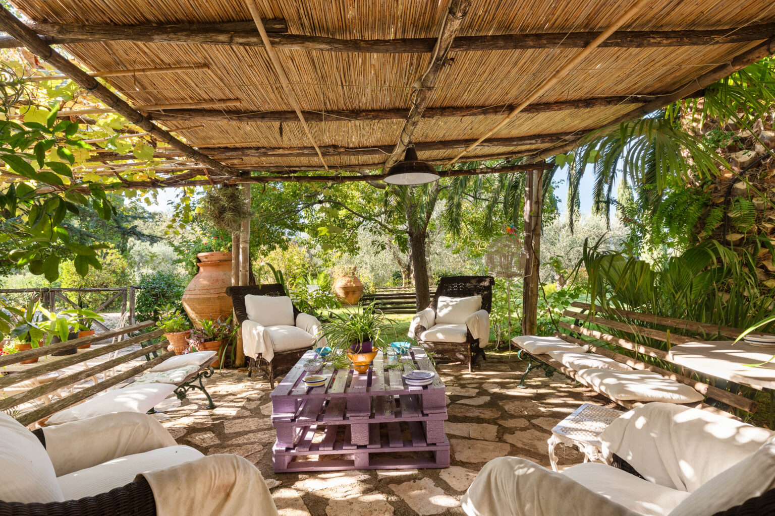 Cozy outdoor seating area under a thatched roof, featuring comfortable chairs and a purple wooden pallet table, surrounded by lush greenery and decorative plants, ideal for relaxation and social gatherings. | Only on SopranoVillas