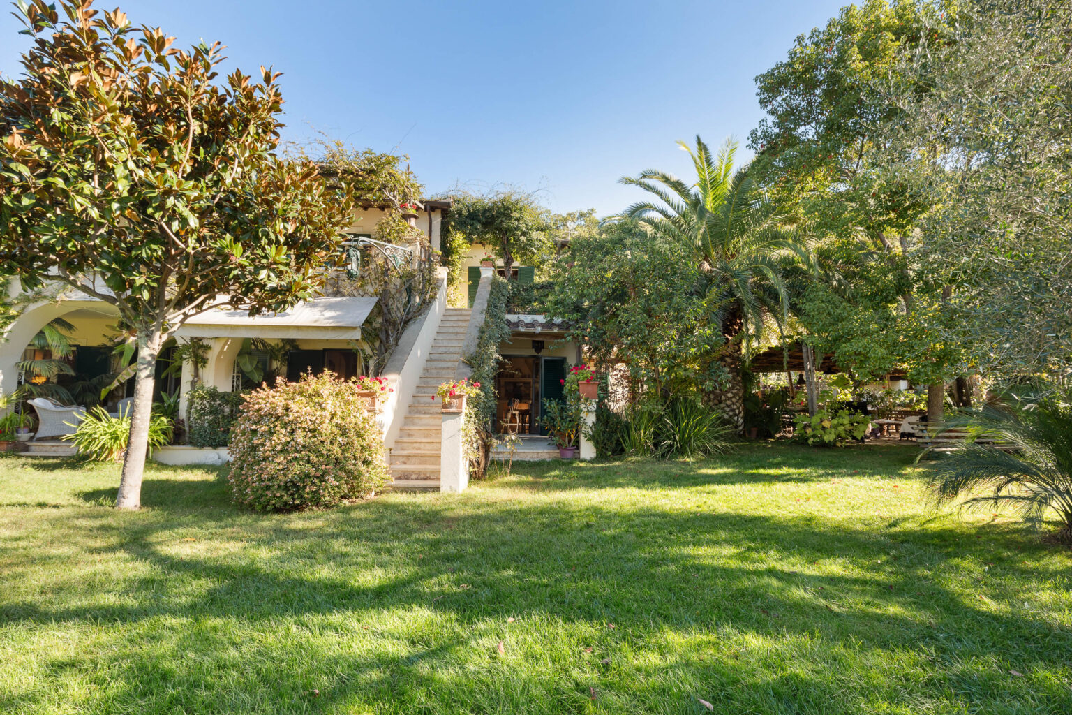 Charming garden view of a two-story house surrounded by lush greenery, featuring a staircase leading to a shaded entrance, vibrant flowers, and palm trees under a clear blue sky. | Only on SopranoVillas