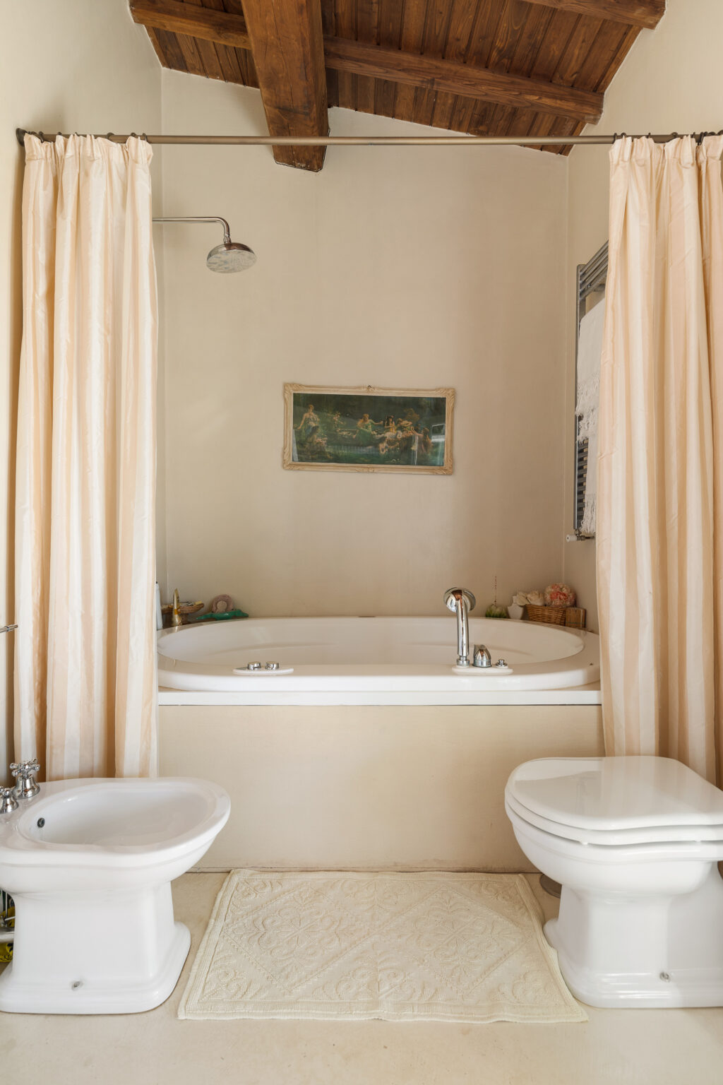 Modern bathroom featuring a white bathtub with a chrome faucet, a shower curtain, a bidet, and a toilet. The space has a warm, inviting atmosphere with wooden beams and soft beige walls, complemented by a decorative wall painting. A plush rug adds comfort to the floor. | Only on SopranoVillas
