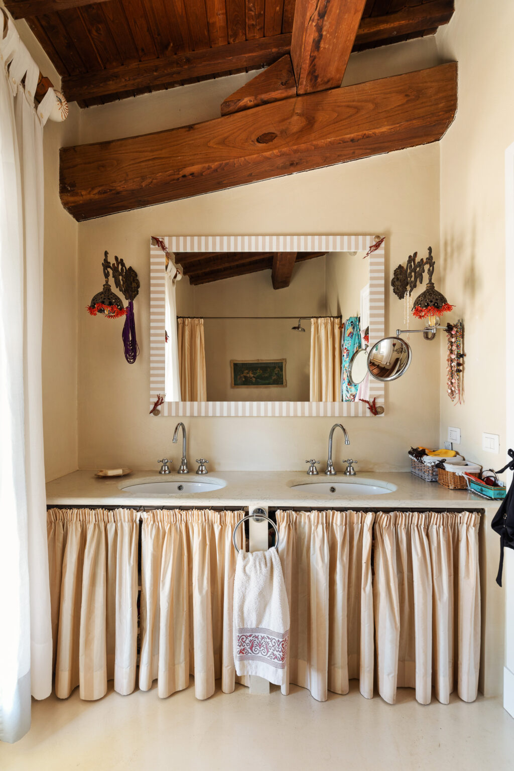 Modern bathroom design featuring a double sink vanity with a concrete countertop, decorative beige curtains, and a large striped mirror. The space includes wooden beams for a rustic touch, along with various decorative elements like wall sconces and accessories. | Only on SopranoVillas