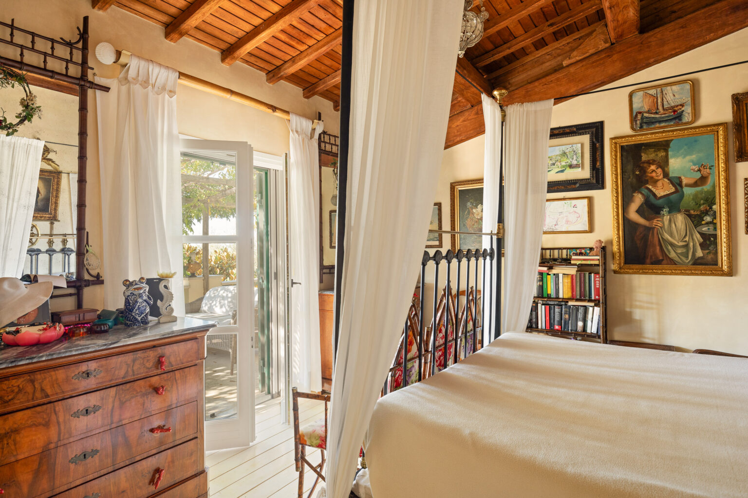 Cozy bedroom interior featuring a four-poster bed, wooden beams, and vintage decor. The room is adorned with framed artwork and a wooden dresser, with sheer curtains allowing natural light to enter from an adjacent balcony. | Only on SopranoVillas