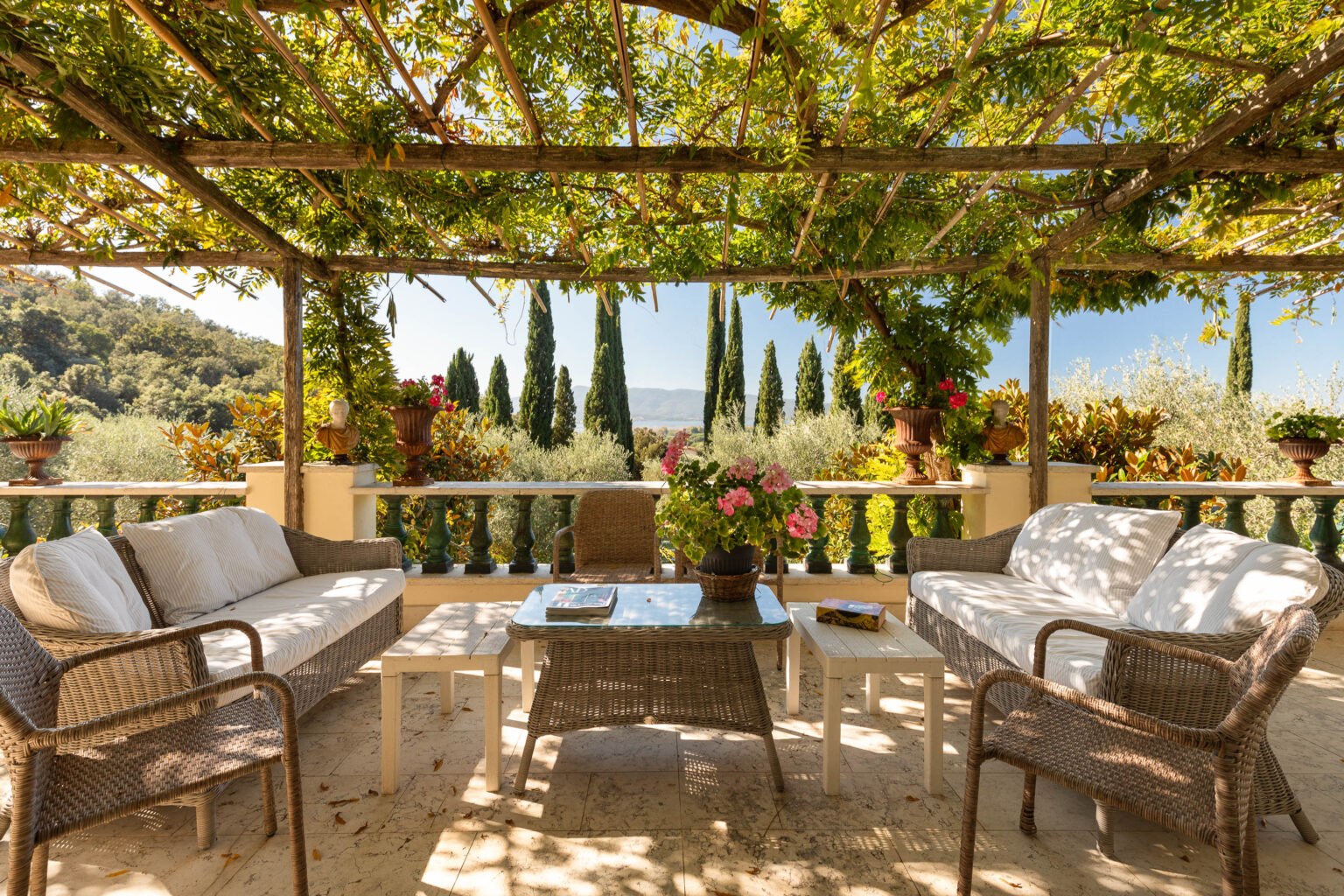 Cozy outdoor seating area under a wooden pergola, surrounded by lush greenery and flowering plants, with a scenic view of tall cypress trees and rolling hills in the background. | Only on SopranoVillas