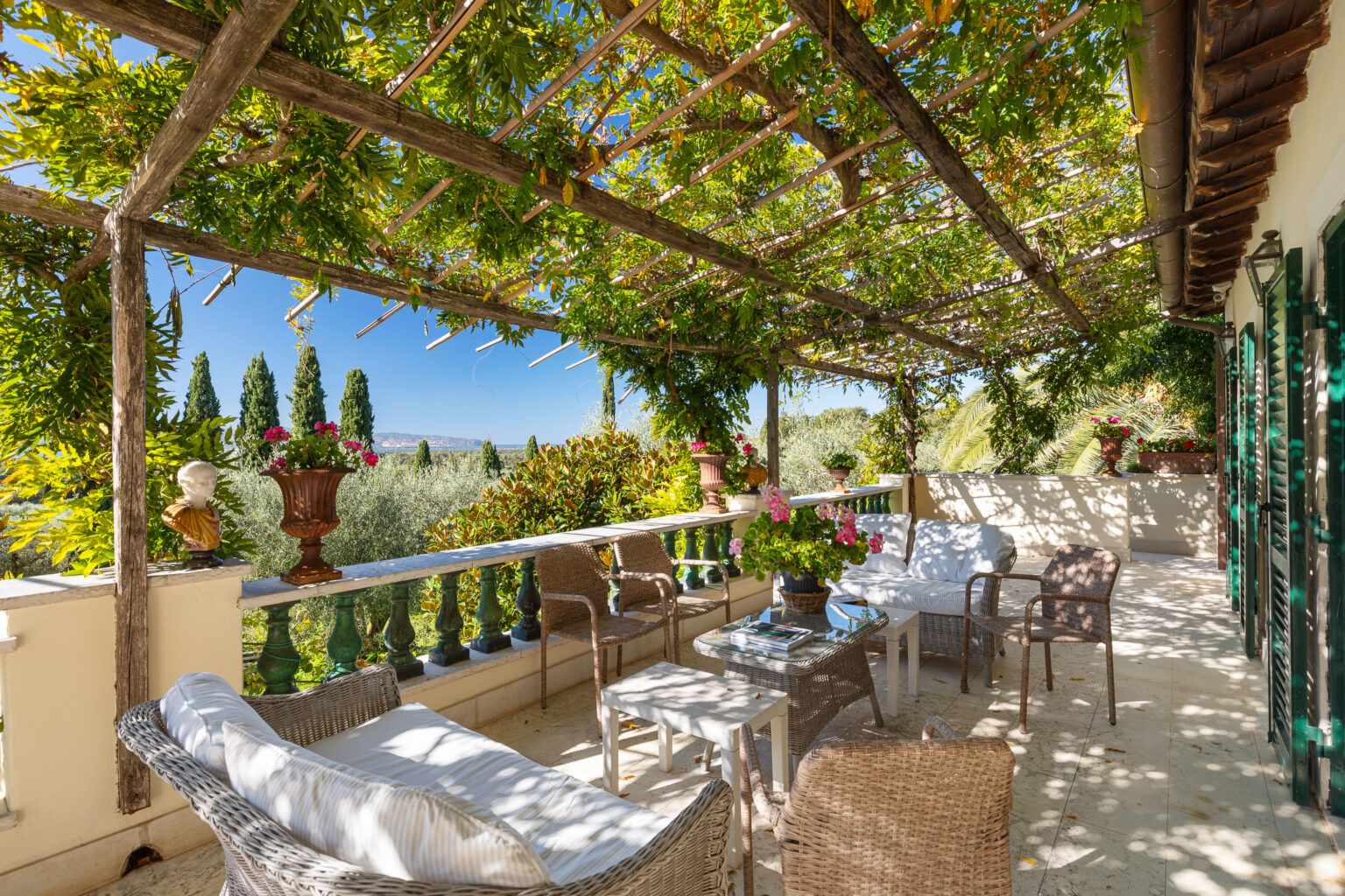 Charming outdoor patio with a wooden pergola covered in greenery, featuring comfortable wicker furniture and vibrant flower pots, overlooking a scenic landscape with cypress trees and blue skies. Perfect for relaxation and enjoying nature. | Only on SopranoVillas