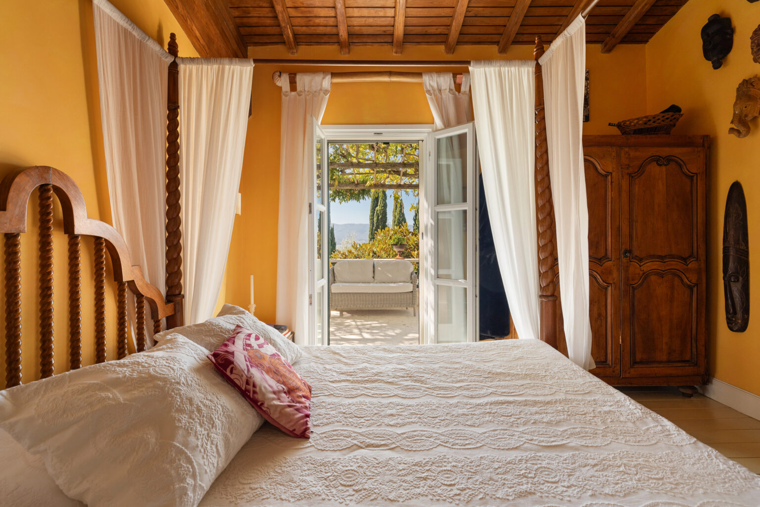 Cozy bedroom featuring a carved wooden bed with decorative pillows, sheer white curtains, and a warm yellow wall. The open double doors lead to a terrace with a comfortable seating area surrounded by greenery, creating a serene indoor-outdoor atmosphere. | Only on SopranoVillas