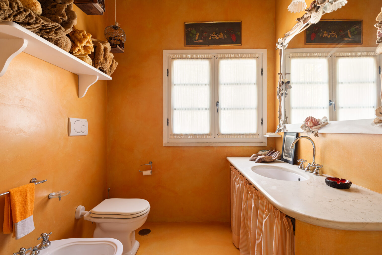 Brightly colored bathroom featuring warm orange walls, a white countertop with a sink, decorative seashells, and a window with sheer curtains. The space includes a toilet, towel rack with orange and gray towels, and a shelf displaying natural sponges, creating a cozy and inviting atmosphere. | Only on SopranoVillas