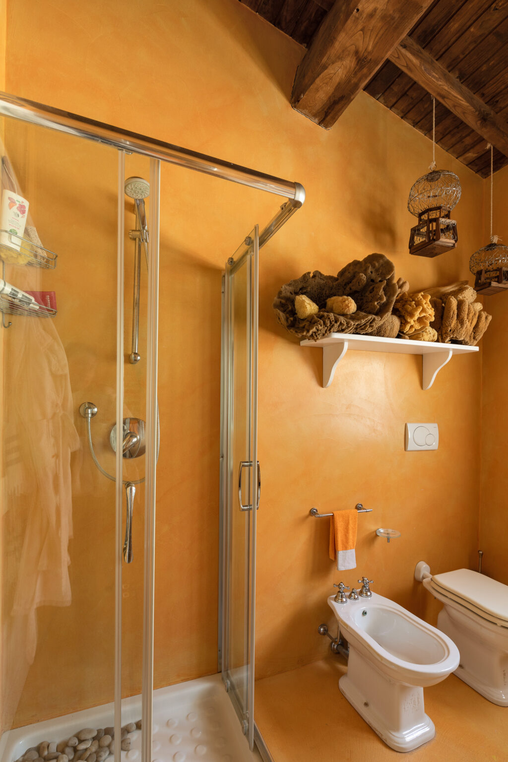 Modern bathroom with a glass shower enclosure, orange textured walls, and decorative natural sponges on a shelf. Features include a bidet, toilet, and towel rack with an orange towel. The wooden ceiling adds warmth to the space. | Only on SopranoVillas
