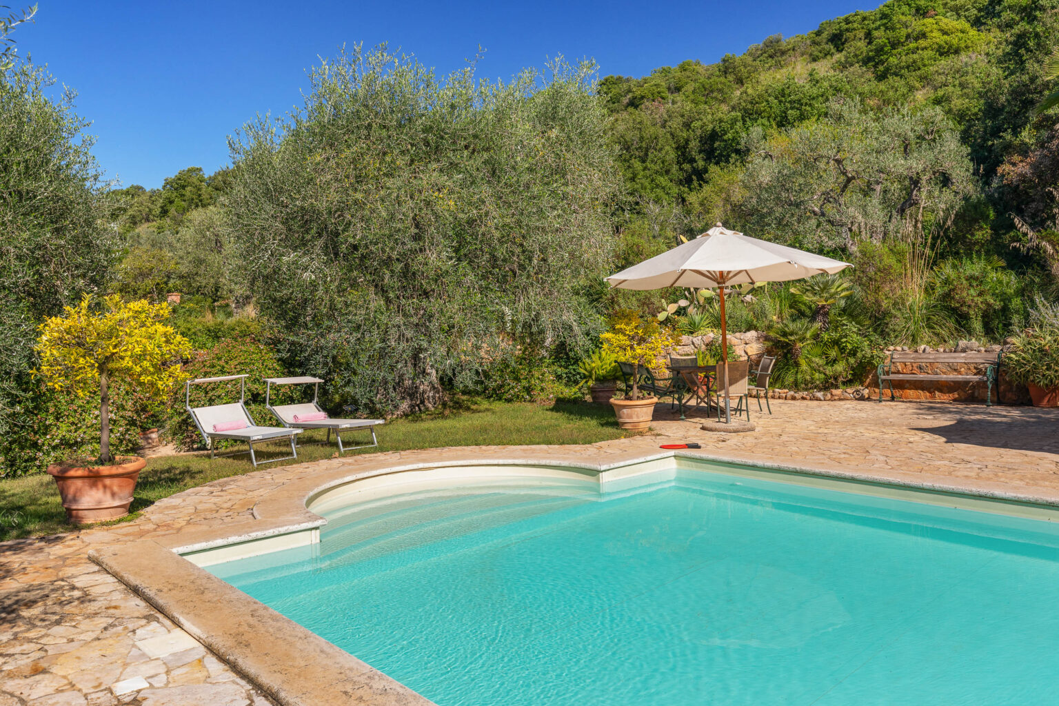 Luxurious outdoor pool area surrounded by lush greenery, featuring two lounge chairs, a shaded table with chairs, and vibrant yellow flowers under a clear blue sky. Ideal for relaxation and leisure. | Only on SopranoVillas