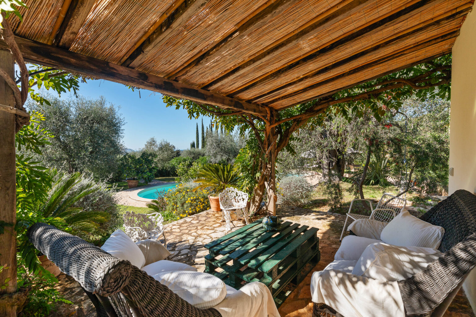 Cozy outdoor seating area with a rustic wooden table and plush cushions, surrounded by lush greenery and a swimming pool in the background, perfect for relaxation and leisure. | Only on SopranoVillas