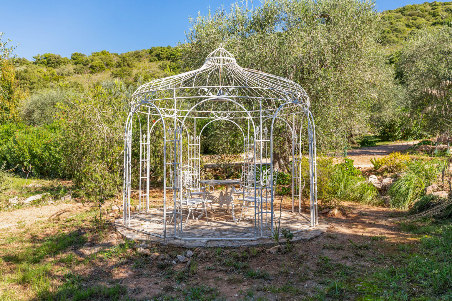 White wrought iron gazebo surrounded by lush greenery, featuring a stone floor and elegant seating, ideal for outdoor relaxation and gatherings. | Only on SopranoVillas