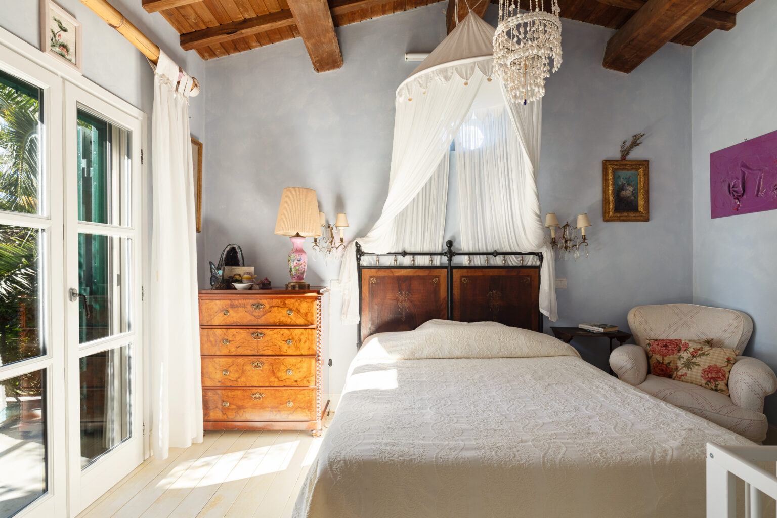 Cozy bedroom featuring a vintage wooden bed with a decorative white coverlet, a stylish chandelier, and a wooden dresser. Natural light streams in through large windows adorned with white curtains, creating a warm and inviting atmosphere. The room is decorated with art and a comfortable armchair, enhancing its charm. | Only on SopranoVillas