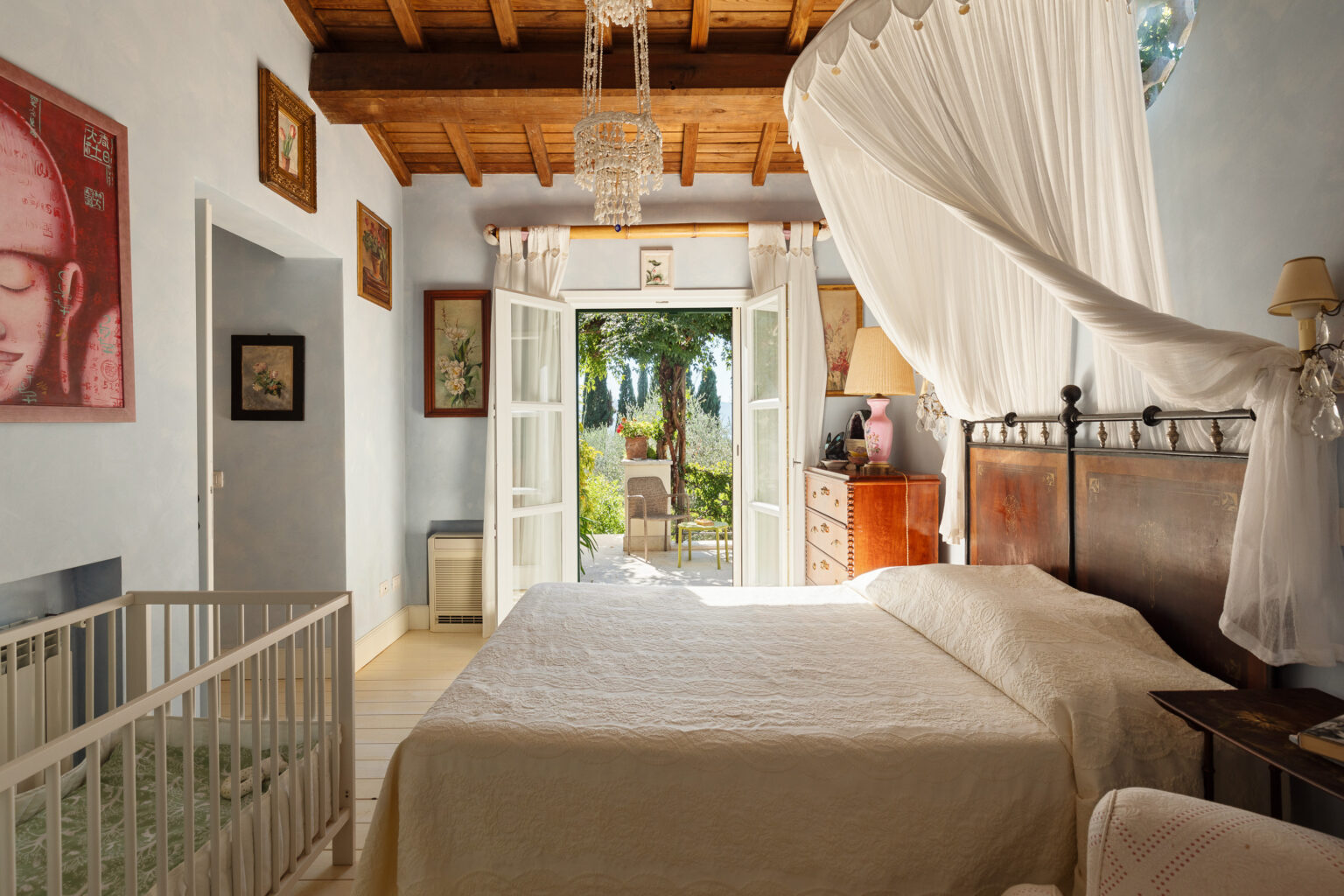 Cozy bedroom with a white quilted bedspread, decorative artwork on the walls, and a crib in the corner. The room features large open doors leading to a garden, allowing natural light to fill the space. A vintage dresser and a pink lamp add charm to the decor, while wooden beams enhance the rustic feel. | Only on SopranoVillas