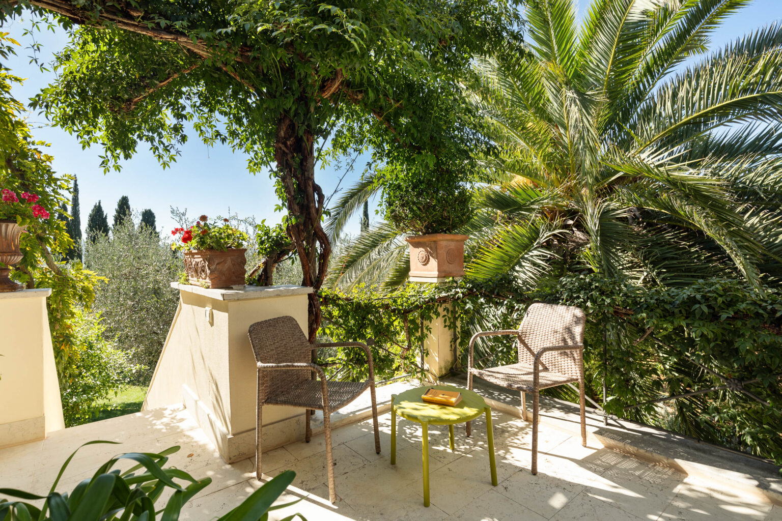 Cozy outdoor seating area featuring two wicker chairs and a round green table, surrounded by lush greenery and flowering plants, under a clear blue sky. Ideal for relaxation and enjoying nature. | Only on SopranoVillas