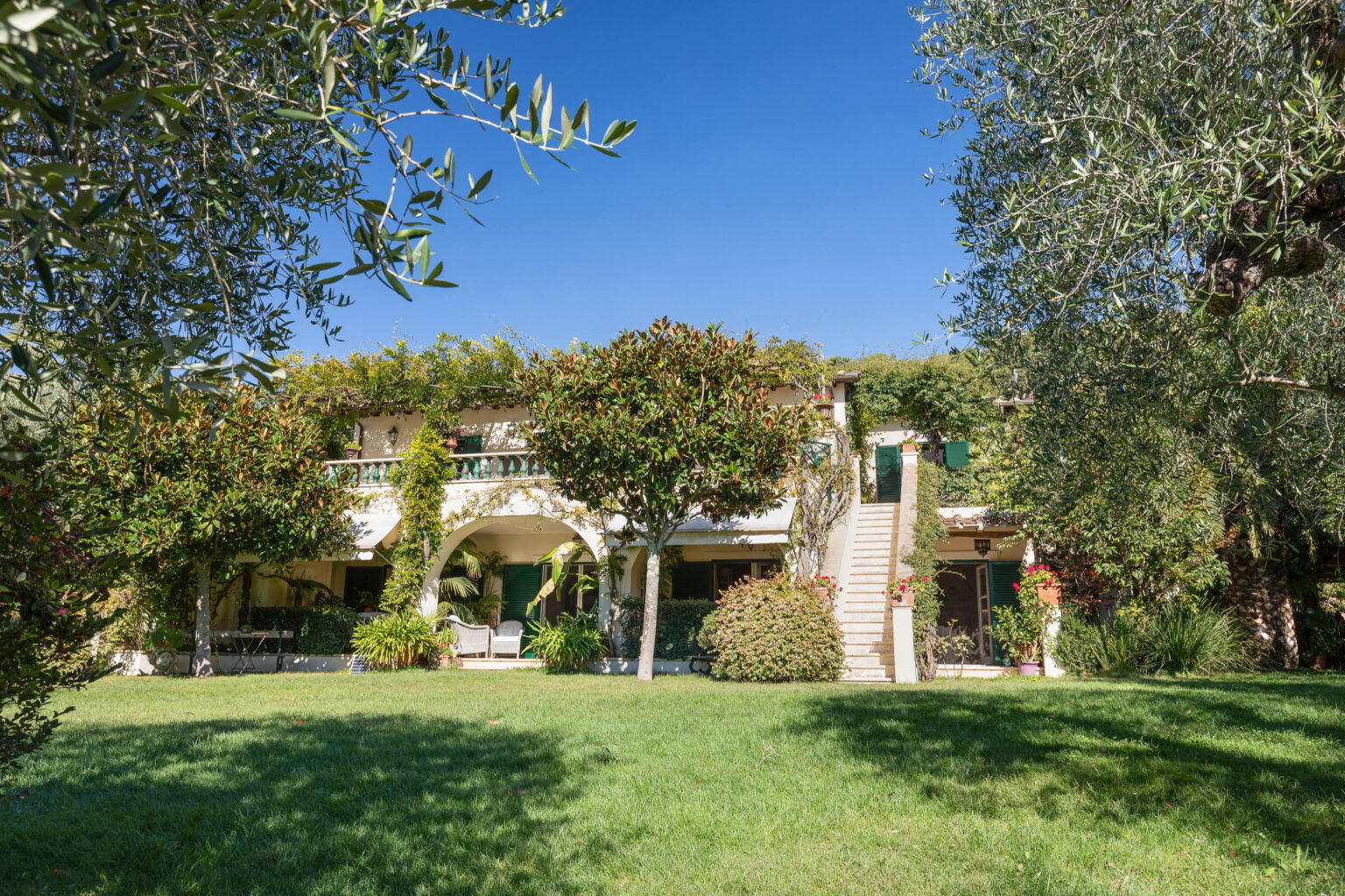 Charming two-story villa surrounded by lush greenery and colorful trees, featuring a spacious lawn, arched entryway, and decorative balcony, set against a clear blue sky. Ideal for outdoor relaxation and picturesque views. | Only on SopranoVillas