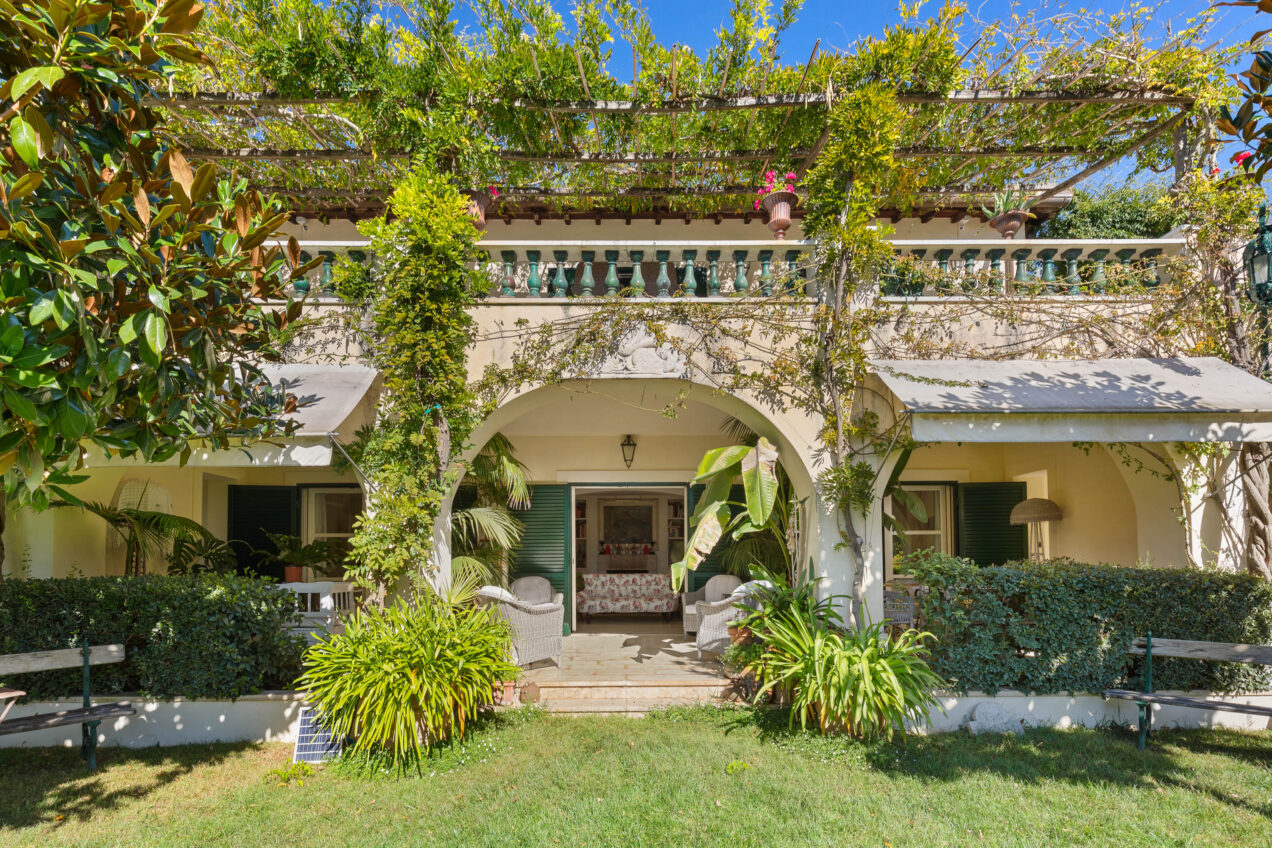 Charming house exterior surrounded by lush greenery, featuring a covered porch with comfortable seating, decorative plants, and a balcony adorned with colorful pots, set against a clear blue sky. | Only on SopranoVillas