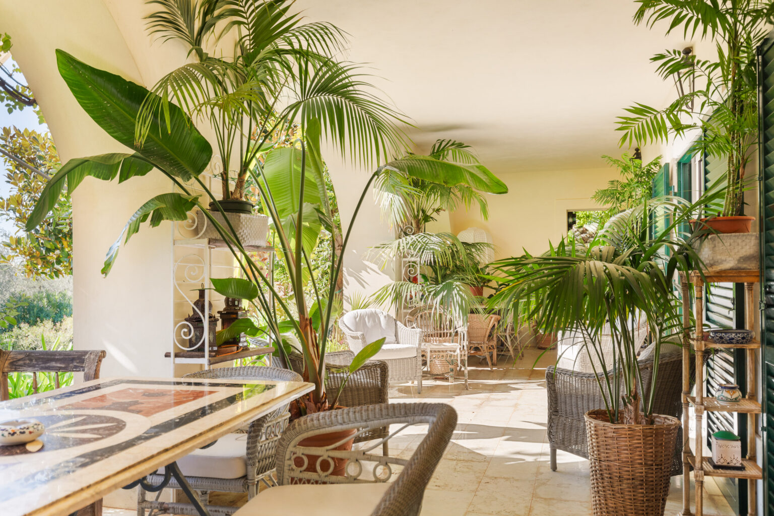 Cozy indoor patio featuring lush green plants, wicker furniture, and a marble table, creating a tranquil and inviting atmosphere for relaxation. Natural light filters through the open space, enhancing the serene environment. | Only on SopranoVillas