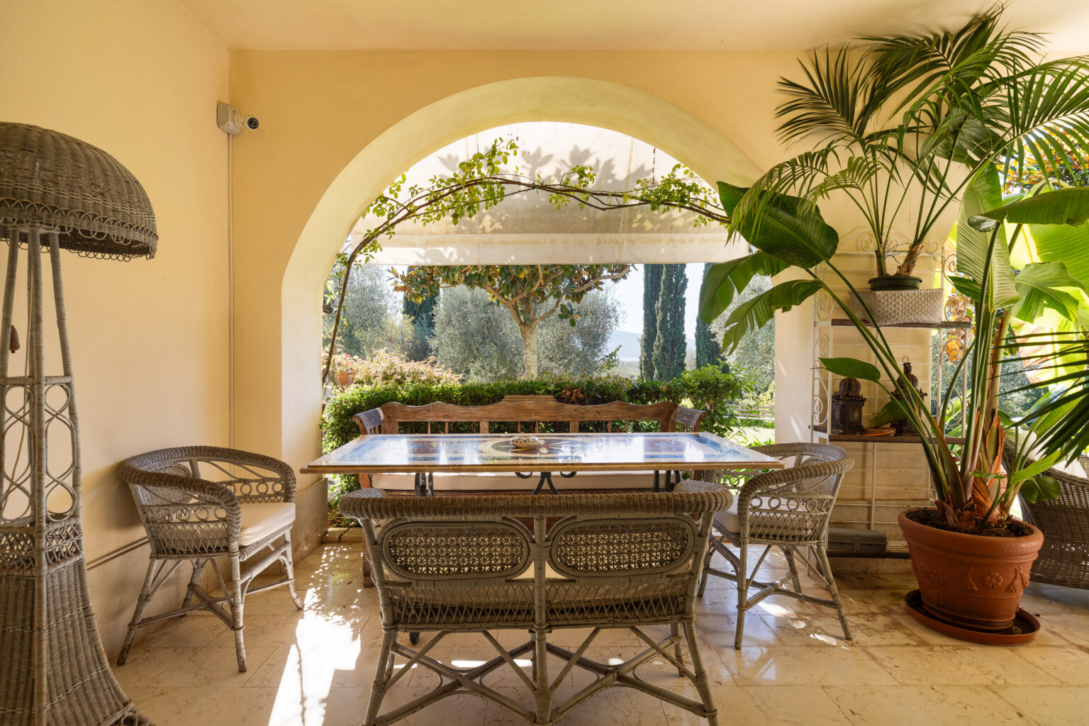 Cozy outdoor dining area featuring a wooden table surrounded by wicker chairs, with lush greenery visible through an arched window. Sunlight filters in, highlighting the elegant design and serene atmosphere. Ideal for relaxing meals or gatherings. | Only on SopranoVillas