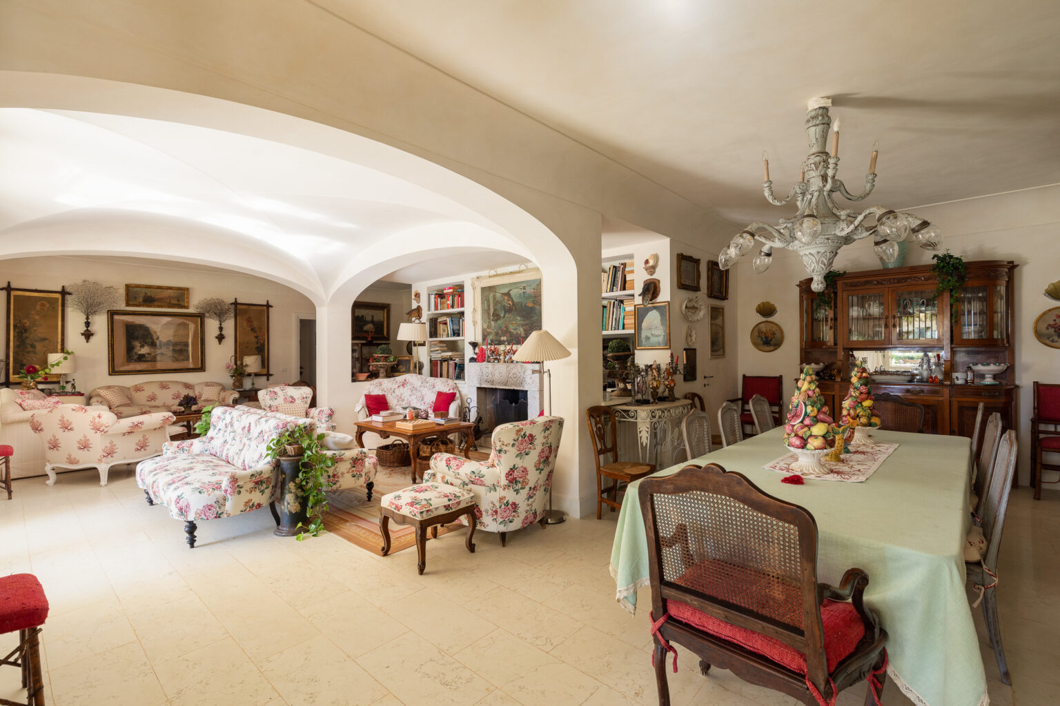 Cozy living room and dining area featuring floral-patterned furniture, a vintage chandelier, and decorative artwork. The space has a warm ambiance with a wooden dining table, ornate chairs, and a bookshelf filled with books and collectibles. | Only on SopranoVillas