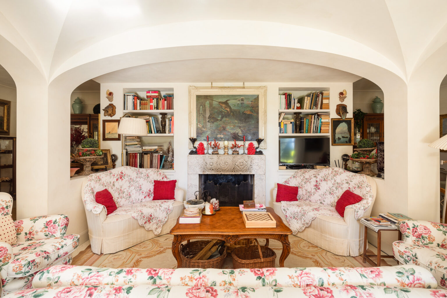 Cozy living room with floral-patterned sofas, a wooden coffee table, and a stone fireplace adorned with decorative items. Bookshelves filled with books and art pieces enhance the inviting atmosphere, complemented by soft lighting and elegant decor. | Only on SopranoVillas