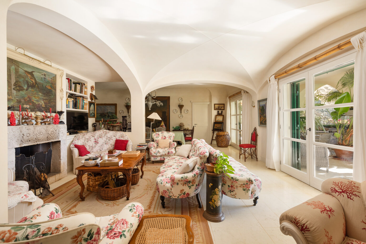 Bright and airy living room featuring floral-patterned sofas, a wooden coffee table, and a decorative stone fireplace. The space is adorned with artwork, bookshelves, and plants, with large windows allowing natural light to fill the room. The elegant arches and soft color palette create a cozy and inviting atmosphere. | Only on SopranoVillas