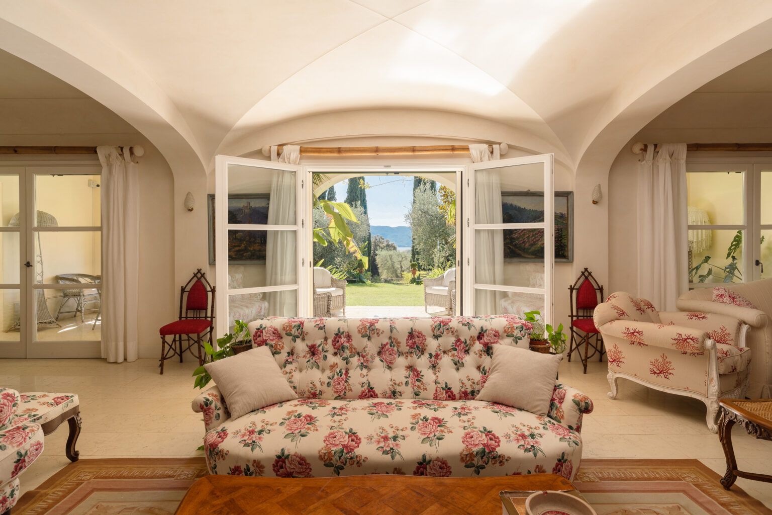 Bright and airy living room featuring a floral-patterned sofa, elegant furnishings, and large open windows that frame a scenic view of a lush garden and distant mountains. Natural light floods the space, enhancing the warm, inviting atmosphere. | Only on SopranoVillas