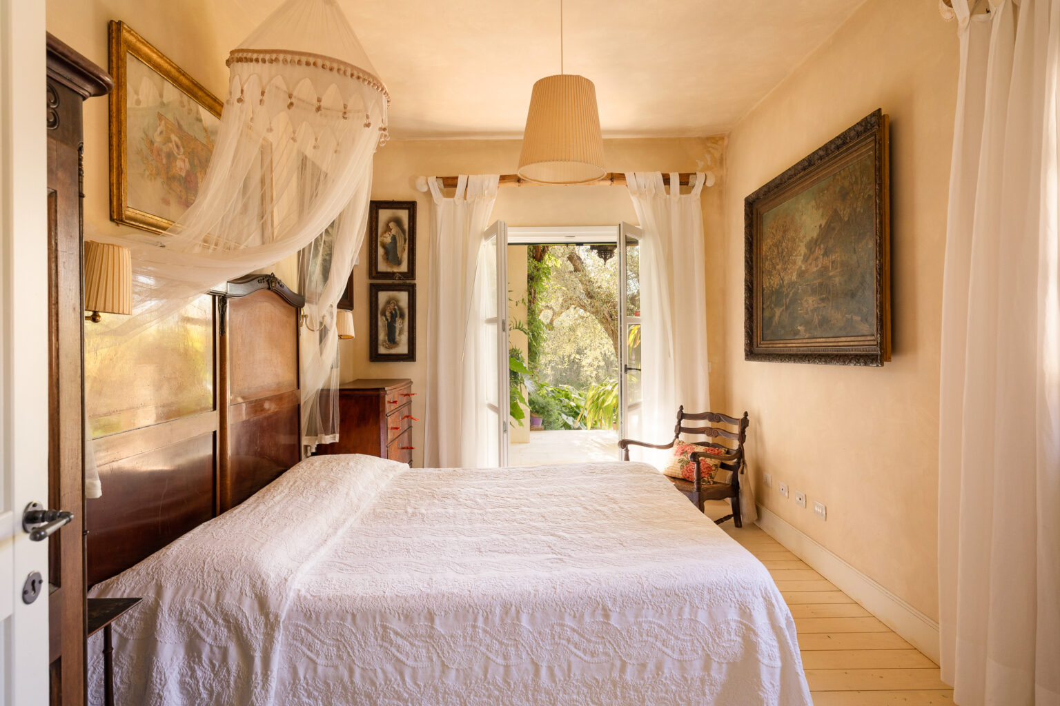 Cozy bedroom featuring a white quilted bedspread, vintage wooden furniture, and soft natural light streaming through sheer curtains. The room includes framed artwork, a rustic chair, and a view of a lush outdoor garden. | Only on SopranoVillas