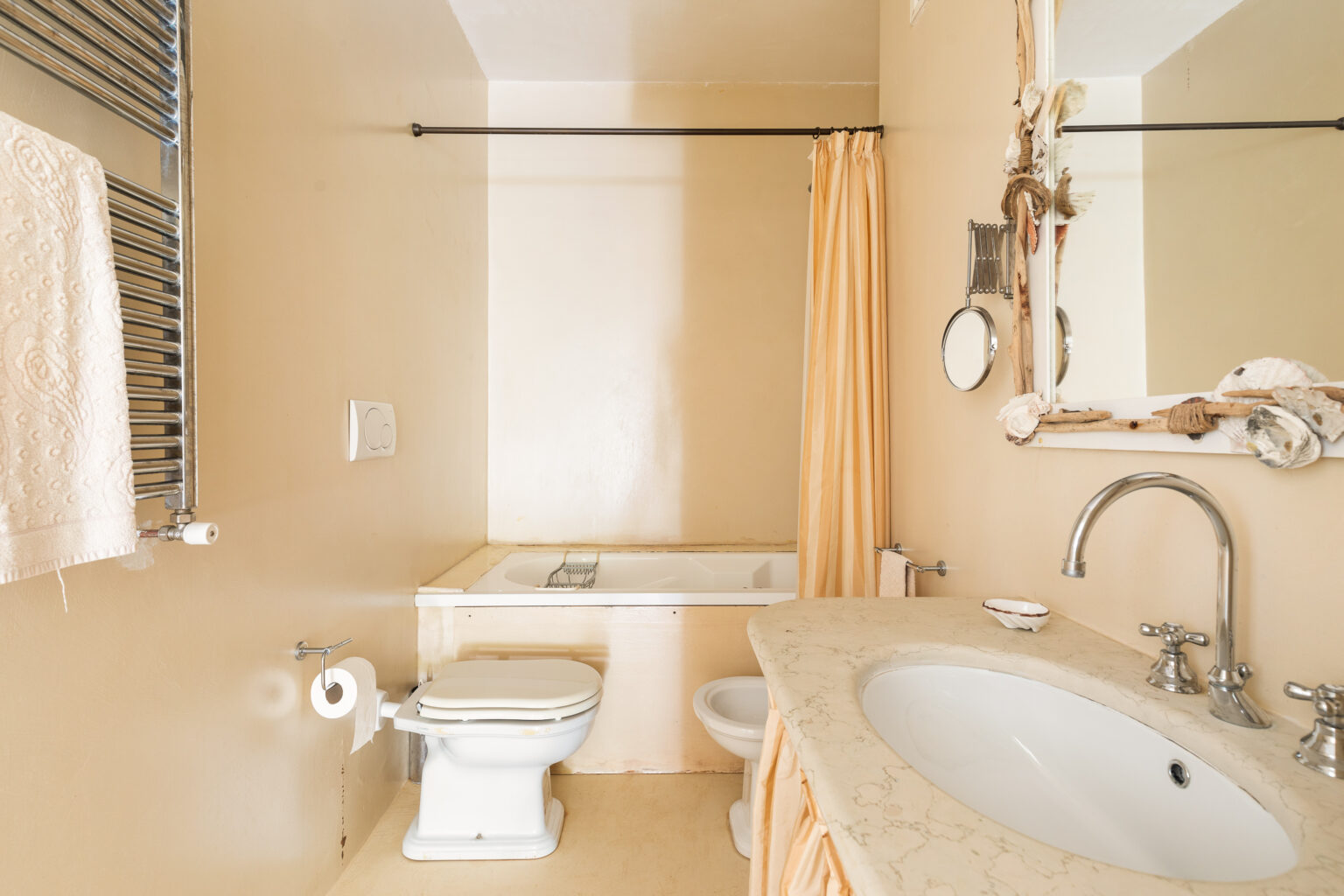 Cozy bathroom featuring a white toilet, a bathtub with a shower curtain, and a stylish marble sink with chrome fixtures. The walls are painted in a soft beige tone, complemented by a towel rack and decorative mirror. Ideal for showcasing modern bathroom design. | Only on SopranoVillas