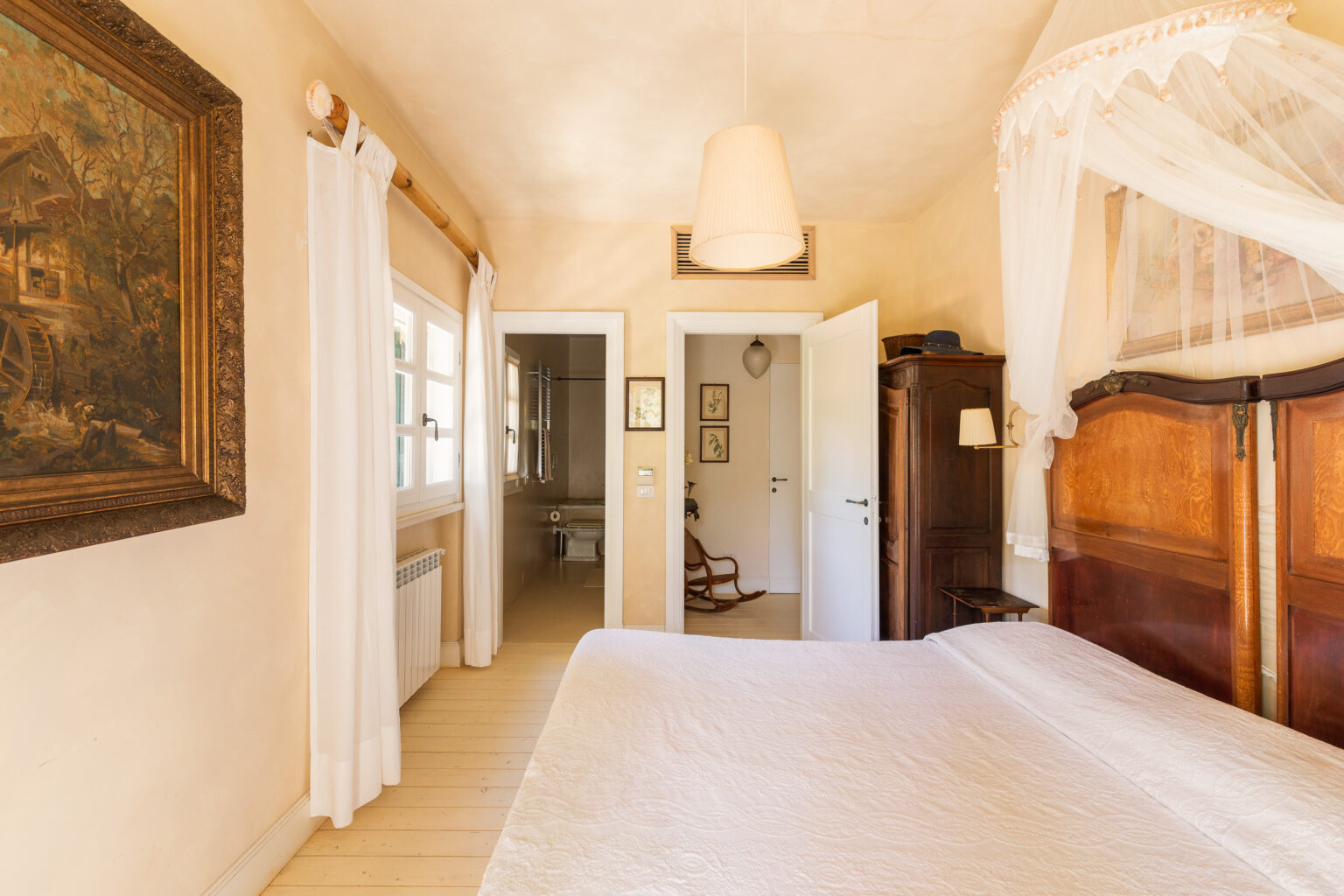 Cozy bedroom featuring a large bed with a white quilt, elegant wooden headboard, and soft lighting. The room includes a vintage painting, sheer white curtains, and a view into an adjoining bathroom. Warm beige walls and natural wood flooring enhance the inviting atmosphere. | Only on SopranoVillas