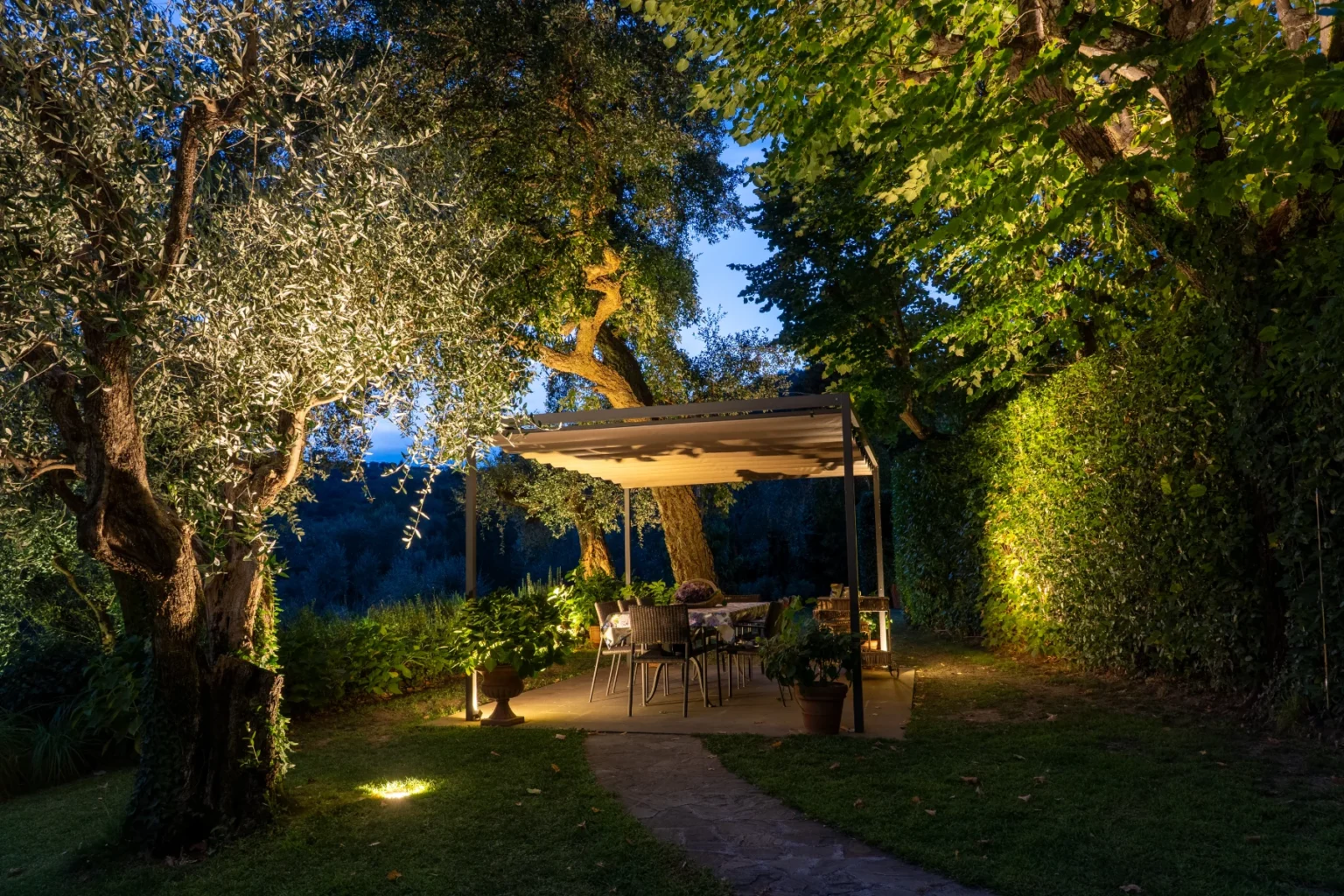 A cozy outdoor dining area illuminated at dusk, featuring a covered patio with a table and chairs surrounded by lush greenery and olive trees. Soft lighting highlights the pathway and plants, creating a serene and inviting atmosphere for evening gatherings. | Only on SopranoVillas