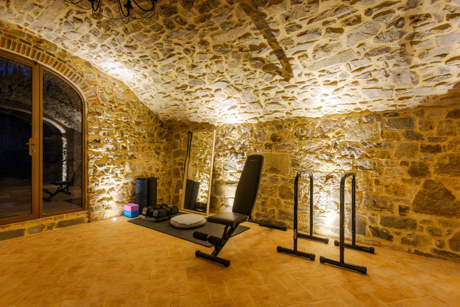 Home gym in a stone-walled basement with workout equipment, including a bench and pull-up bars, illuminated by soft lighting. | Only on SopranoVillas