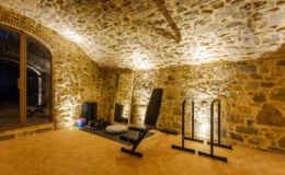 Home gym in a stone-walled basement with workout equipment, including a bench and pull-up bars, illuminated by soft lighting.