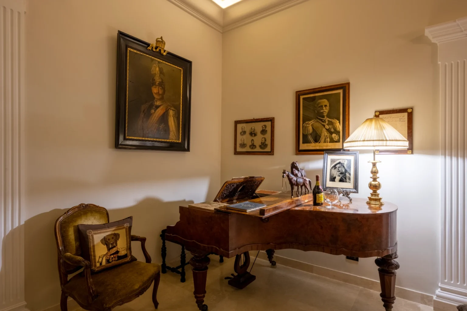 Elegant corner of a vintage room featuring a polished wooden piano, a decorative lamp, and framed historical portraits. A plush chair with a dog-themed cushion adds charm, while a small horse figurine and wine bottle complete the sophisticated decor. | Only on SopranoVillas