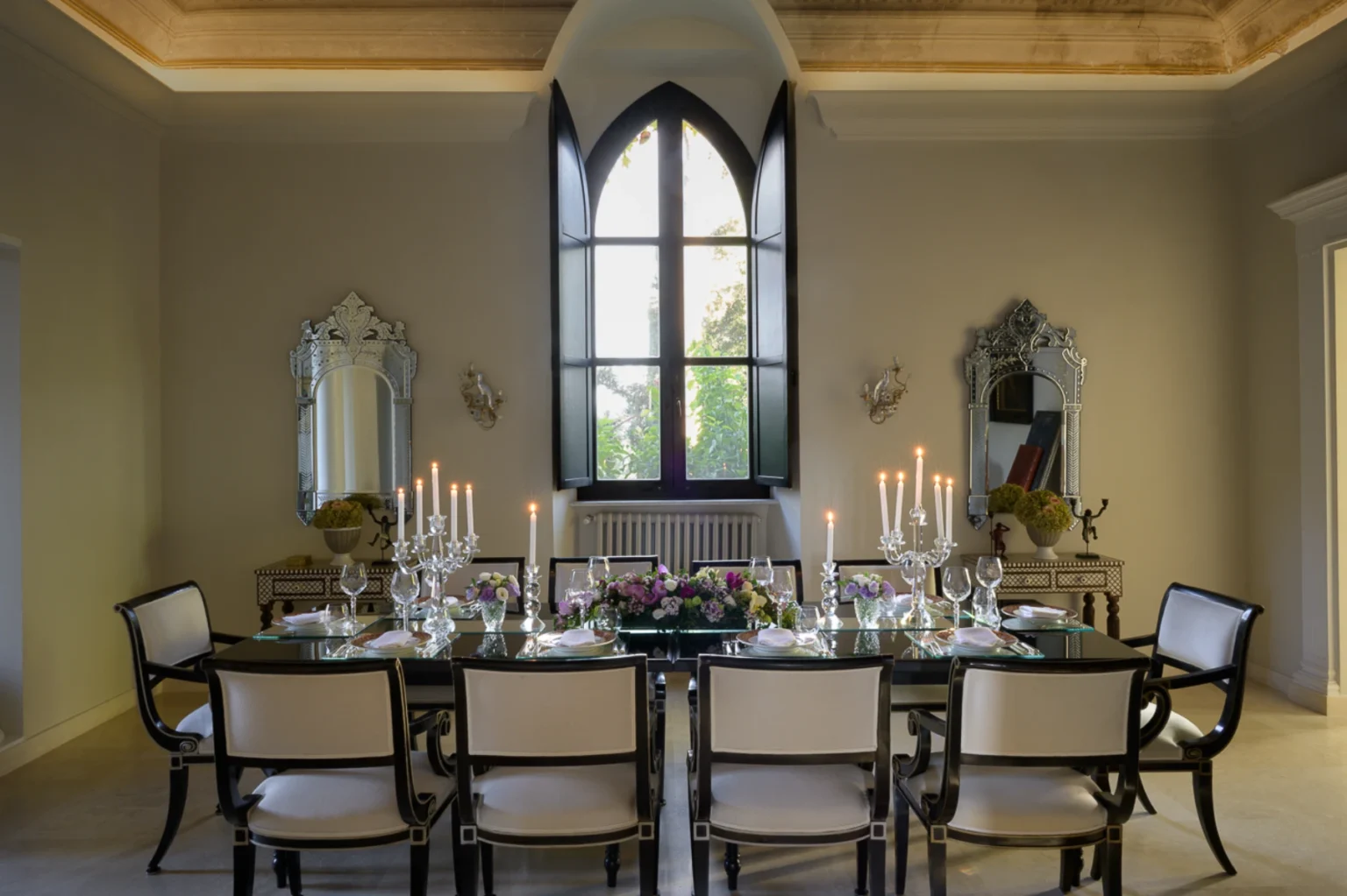 Elegant dining room featuring a long glass table set for a formal meal, surrounded by stylish black and white chairs. The room is illuminated by candles in crystal holders and adorned with floral centerpieces. Large gothic windows provide natural light and showcase a view of greenery outside, while ornate mirrors enhance the luxurious ambiance. | Only on SopranoVillas
