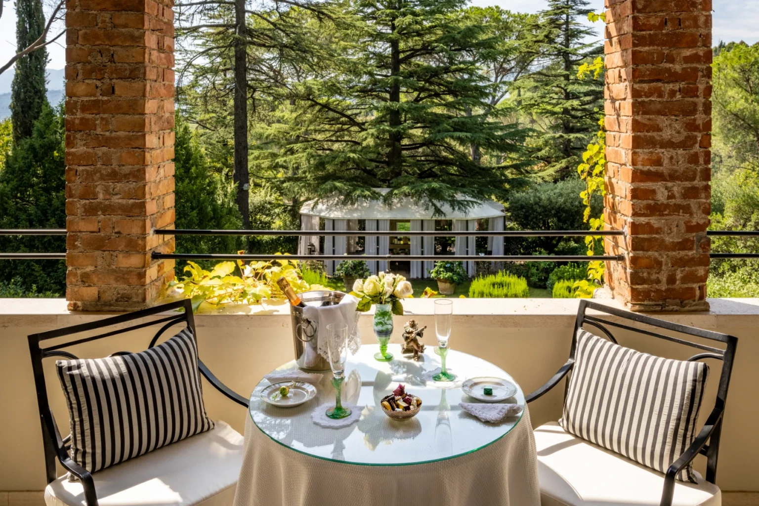 Elegant outdoor dining setup on a balcony featuring a glass table adorned with gourmet treats, champagne, and fresh flowers, surrounded by lush greenery and a charming gazebo in the background. Perfect for a serene dining experience. | Only on SopranoVillas