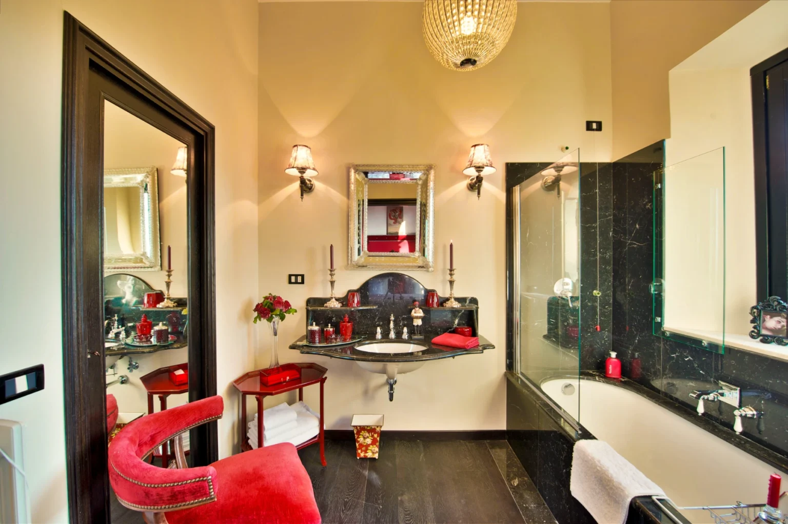 Luxurious bathroom interior featuring elegant design elements such as a marble sink, decorative mirrors, and stylish lighting. The space includes a red velvet chair, a glass shower enclosure, and tasteful decor accents like candles and flowers, creating a sophisticated ambiance. | Only on SopranoVillas