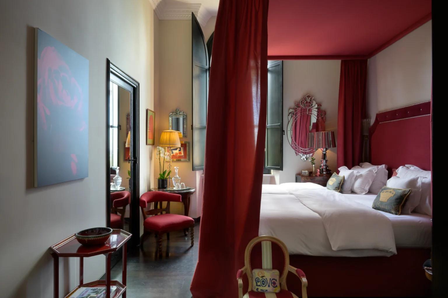 Cozy hotel room featuring a red color scheme, including a plush bed with decorative pillows, elegant furnishings, and large windows that allow natural light. The room is accented with artwork, a stylish mirror, and a charming seating area, creating an inviting atmosphere for guests. | Only on SopranoVillas