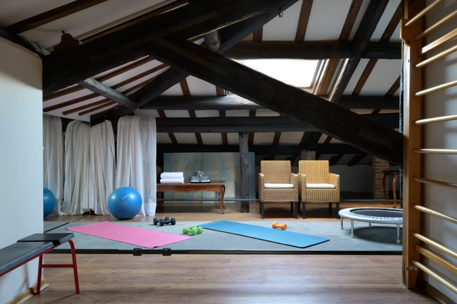 Home gym in an attic space featuring wooden beams, exercise mats, dumbbells, a stability ball, and a trampoline, with a cozy seating area in the background. | Only on SopranoVillas