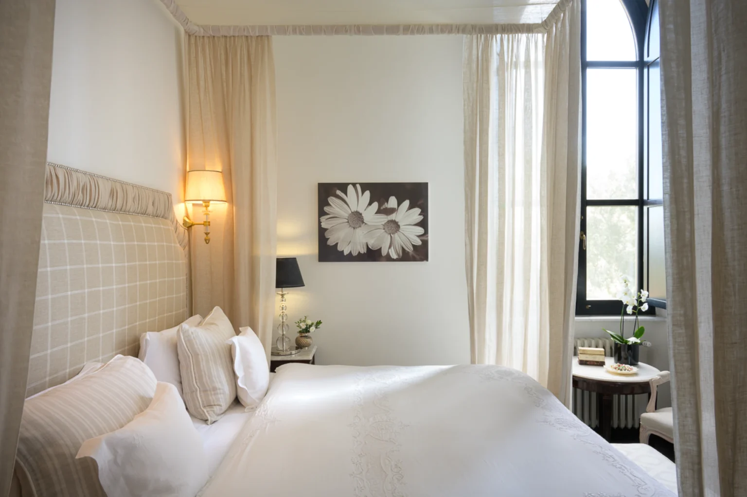 Cozy and elegant bedroom featuring a plush bed with decorative pillows, soft beige curtains, and stylish wall art of daisies. The room is well-lit by a combination of wall sconces and natural light from a large window, enhancing the serene atmosphere. A small side table holds decorative items and a potted plant, adding a touch of nature to the interior design. | Only on SopranoVillas