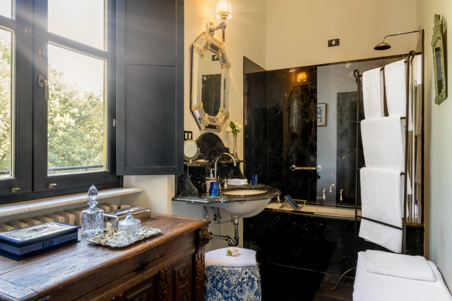 Elegant bathroom interior featuring a black marble sink, ornate mirror, and a towel rack. Natural light streams through large windows, highlighting decorative elements like a wooden chest and glass containers. The space combines modern and classic design elements for a luxurious feel. | Only on SopranoVillas