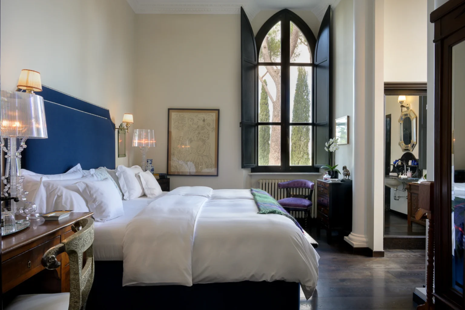 Elegant hotel room featuring a plush bed with white linens, a stylish blue headboard, and decorative lamps. The room includes a large, arched window with a view of greenery, a vintage wooden desk, and a cozy chair. A modern bathroom area is visible, showcasing tasteful decor and amenities. Ideal for luxury travel and relaxation. | Only on SopranoVillas