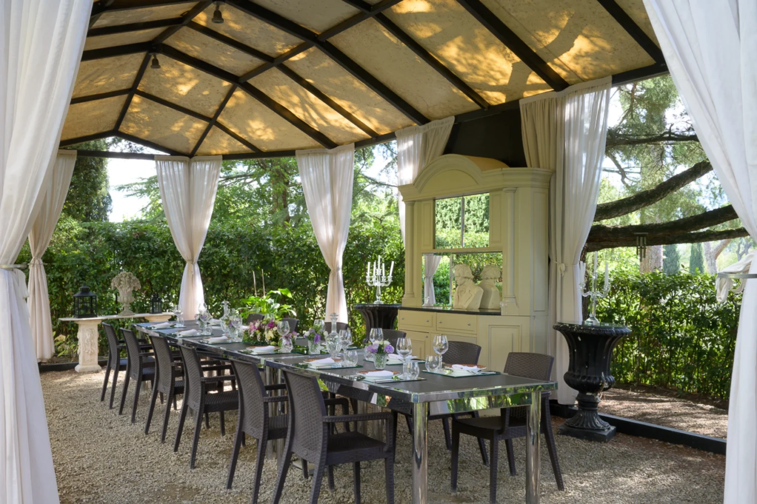 Elegant outdoor dining area under a stylish canopy, featuring a long glass table set with fine china and floral centerpieces, surrounded by plush chairs and lush greenery. Ideal for gatherings and special occasions. | Only on SopranoVillas