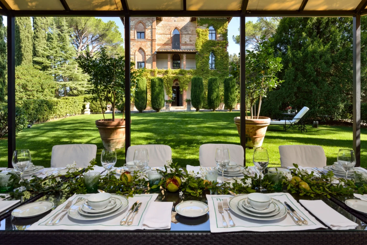 Elegant outdoor dining setup with a beautifully arranged table featuring fine china and glassware, overlooking a manicured garden and a historic villa adorned with greenery. Perfect for al fresco dining experiences. | Only on SopranoVillas