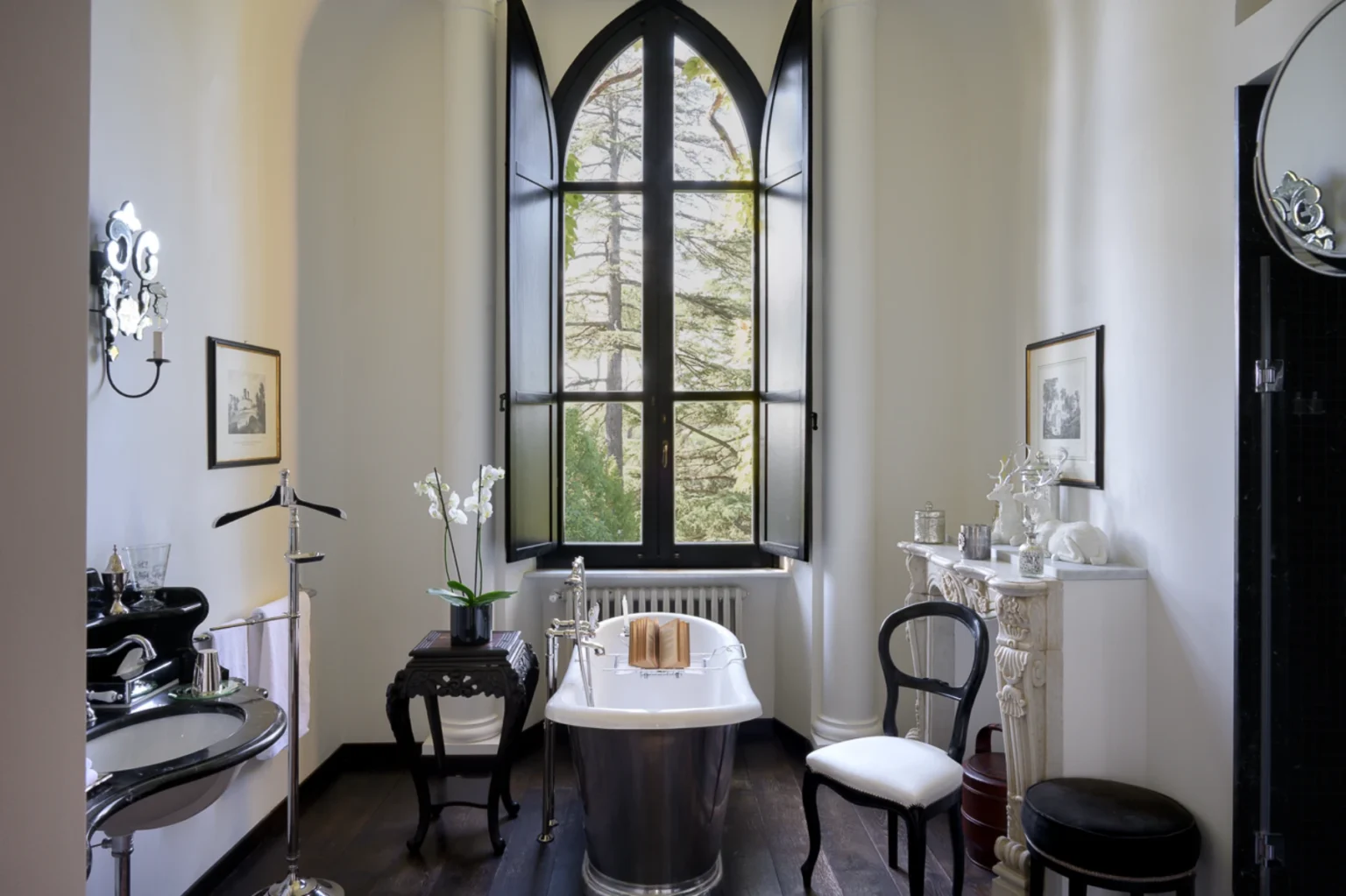 Elegant bathroom featuring a freestanding bathtub, vintage decor, and large gothic-style windows that provide natural light and a view of greenery. The space includes stylish furnishings like a black chair and intricately designed vanity, enhancing its luxurious ambiance. | Only on SopranoVillas