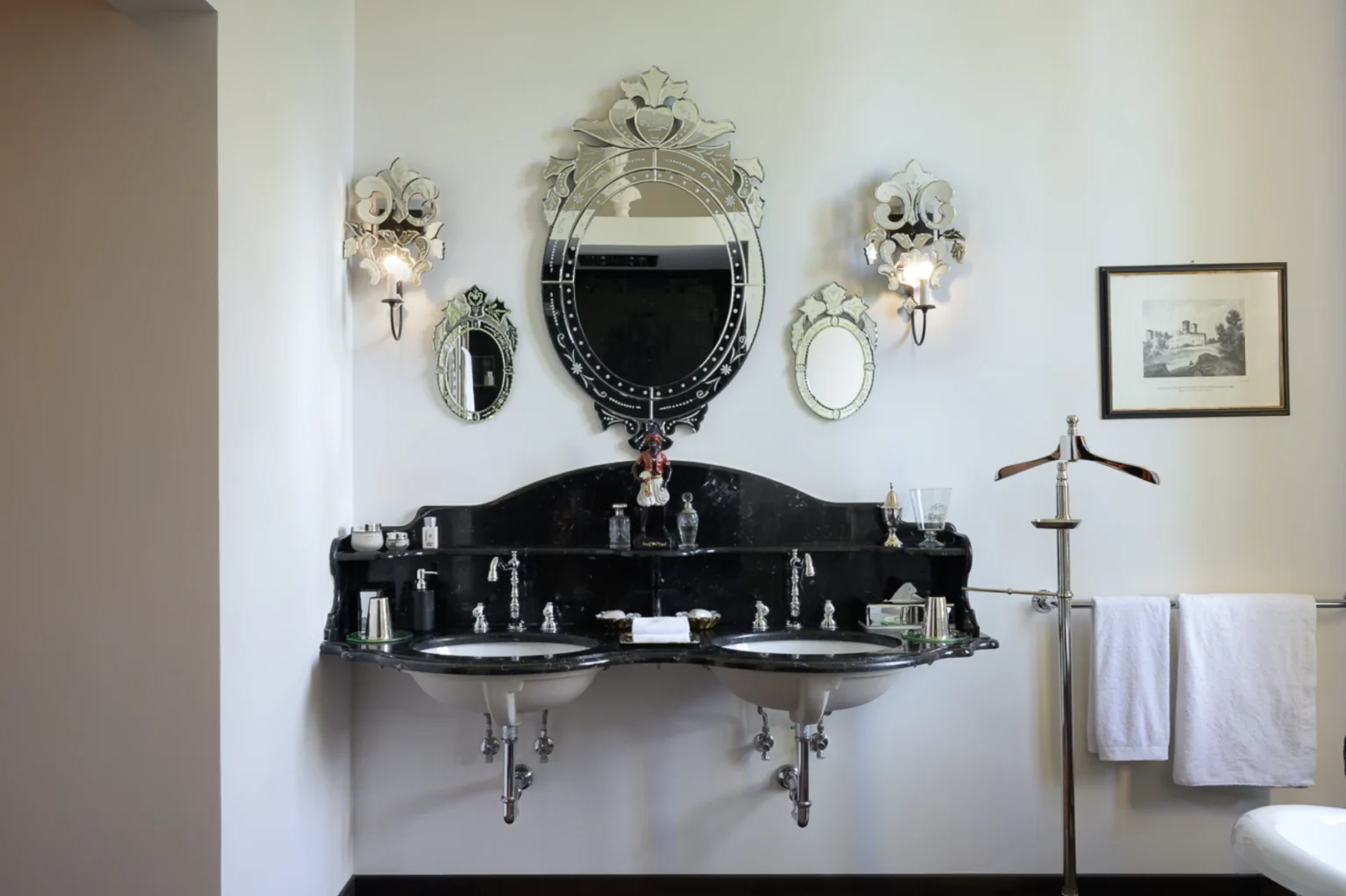 Elegant bathroom featuring a double sink vanity with a black marble countertop, ornate mirrors, and vintage-inspired wall sconces. The decor includes a decorative figurine and neatly arranged toiletries, enhancing the luxurious ambiance. White towels hang on a stylish towel rack, completing the sophisticated look. | Only on SopranoVillas