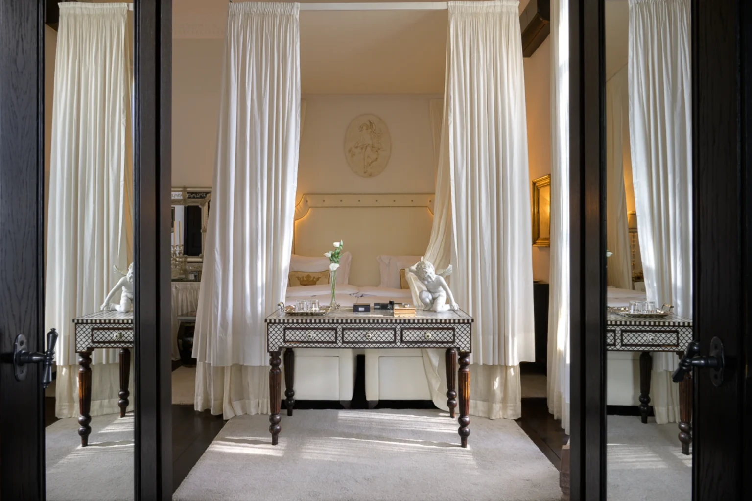 Elegant bedroom interior featuring a four-poster bed with flowing white drapes, a decorative table with a statue, and soft lighting creating a serene atmosphere. The image showcases luxurious decor and a cozy ambiance, perfect for relaxation. | Only on SopranoVillas