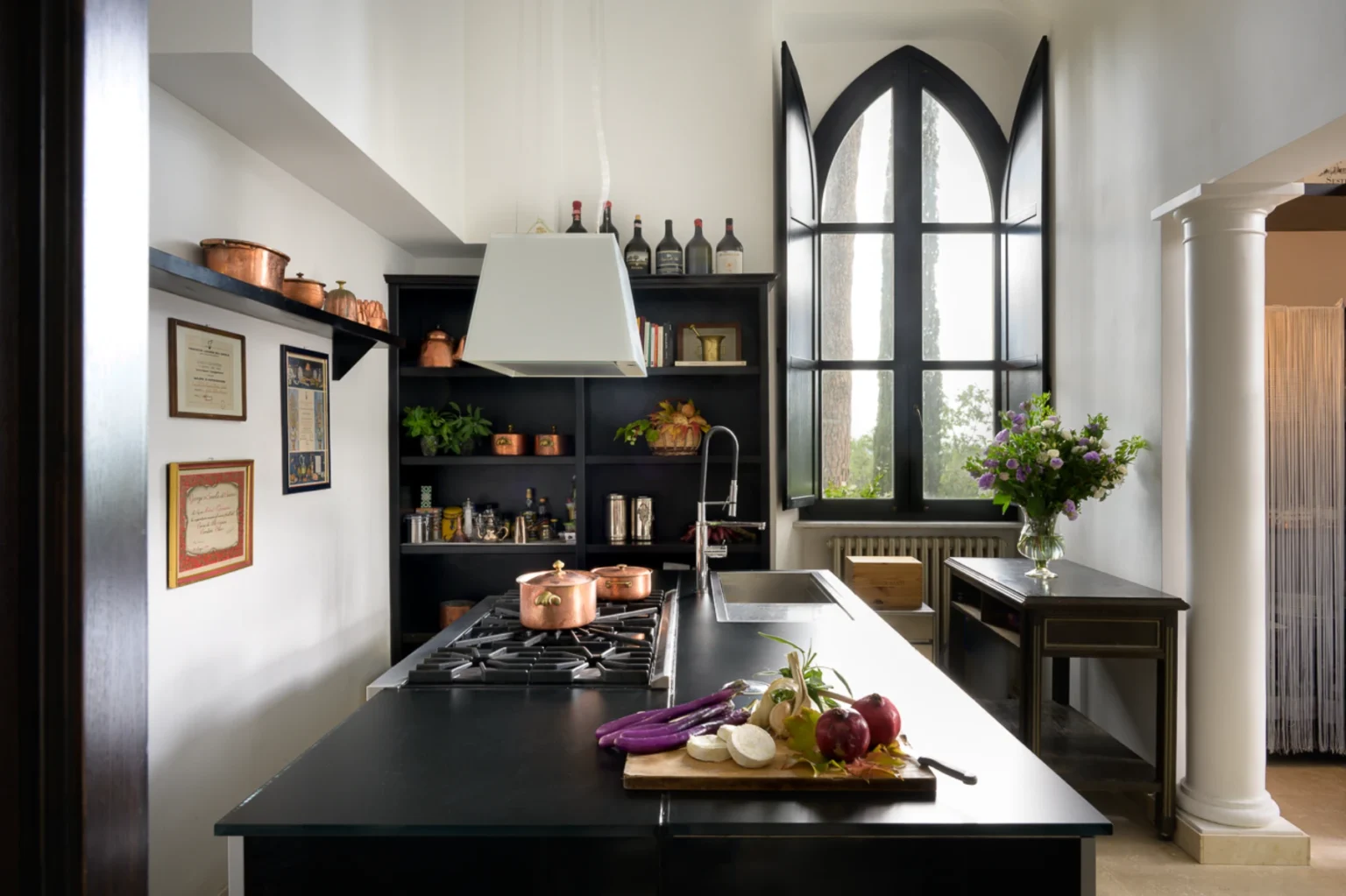 Modern kitchen interior featuring a central island with a gas stove, copper cookware on shelves, and a large gothic-style window allowing natural light. Fresh vegetables and herbs are displayed on the countertop, enhancing the inviting atmosphere. | Only on SopranoVillas